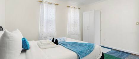 A neatly made bed with white bedding and a blue throw blanket, a white wardrobe, and a patterned carpet.