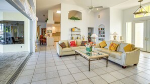 A spacious living room with a large sofa, a coffee table, and a dining area with chairs. There is a kitchen in the background and a view of the outdoors through a glass door. - Pool, Spa & Dock: Waterfront Tarpon Point Home (Cape Coral)