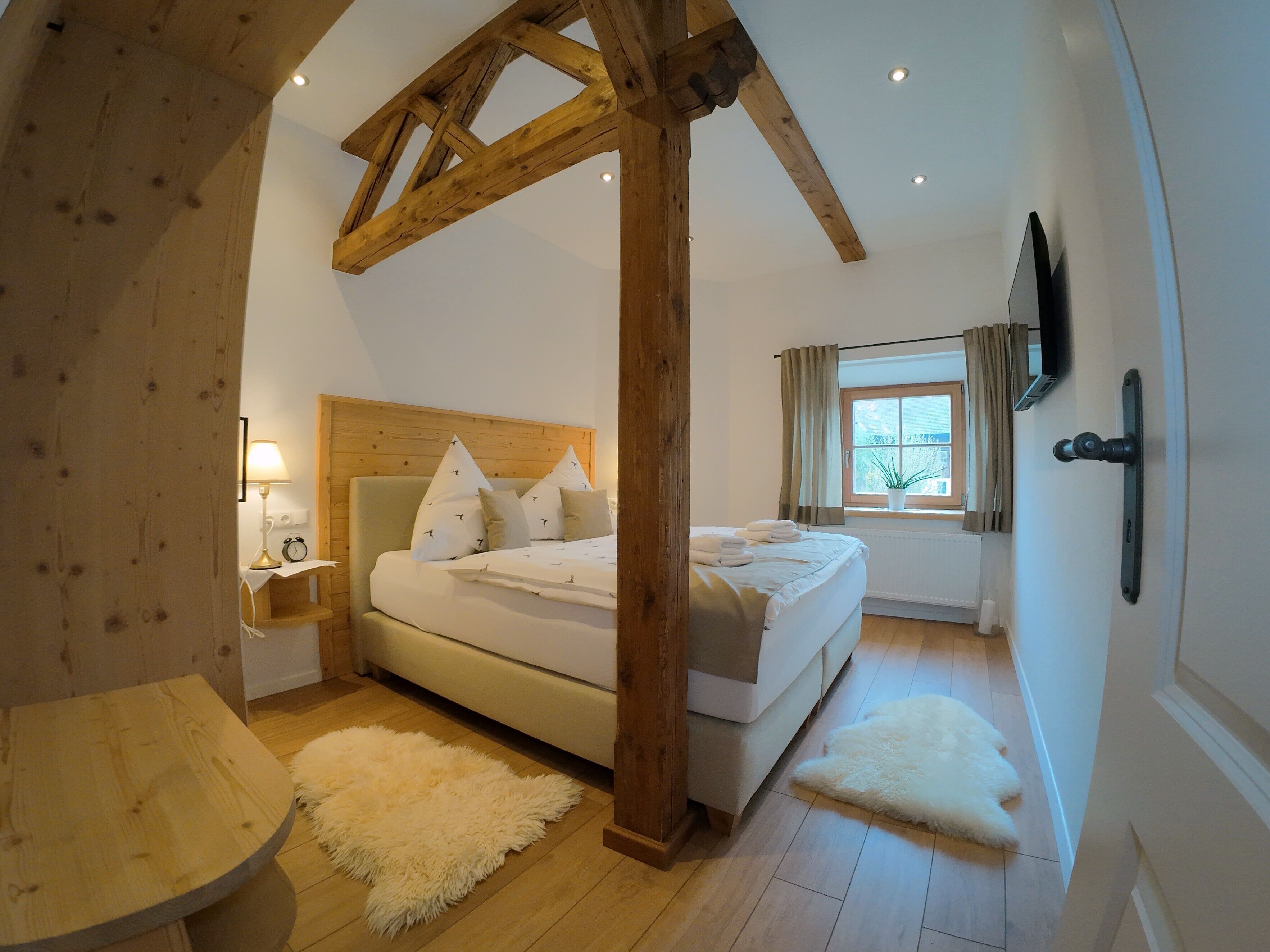 A bedroom with a wooden ceiling, a bed with white bedding, a bedside table with a lamp, a window with curtains, and a white rug.
