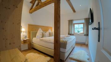 A bedroom with a wooden ceiling, a bed with white bedding, a bedside table with a lamp, a window with curtains, and a white rug.