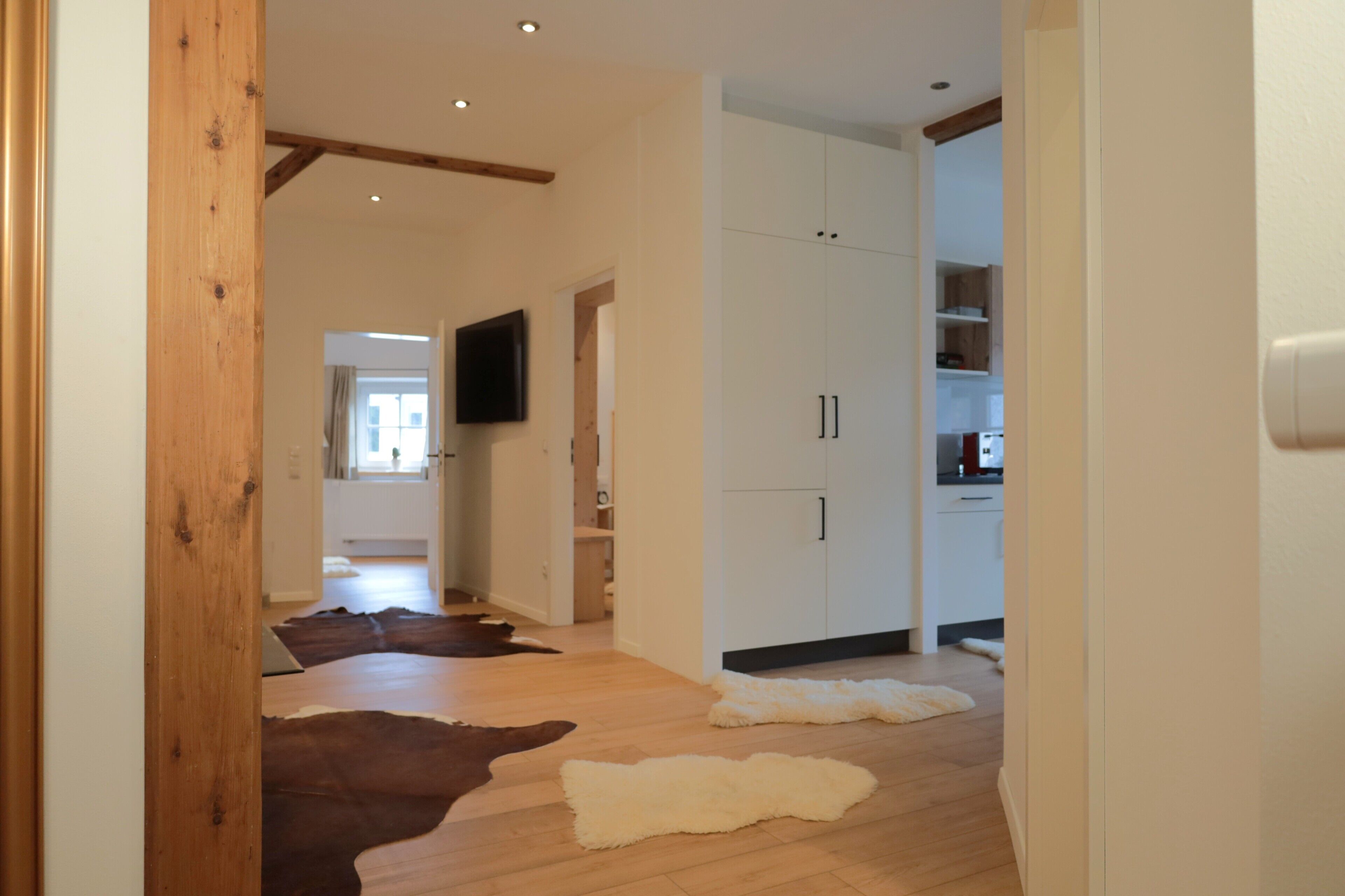 A hallway with wooden flooring, a television mounted on the wall, and a kitchen area visible through an open door.