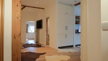 A hallway with wooden flooring, a television mounted on the wall, and a kitchen area visible through an open door.