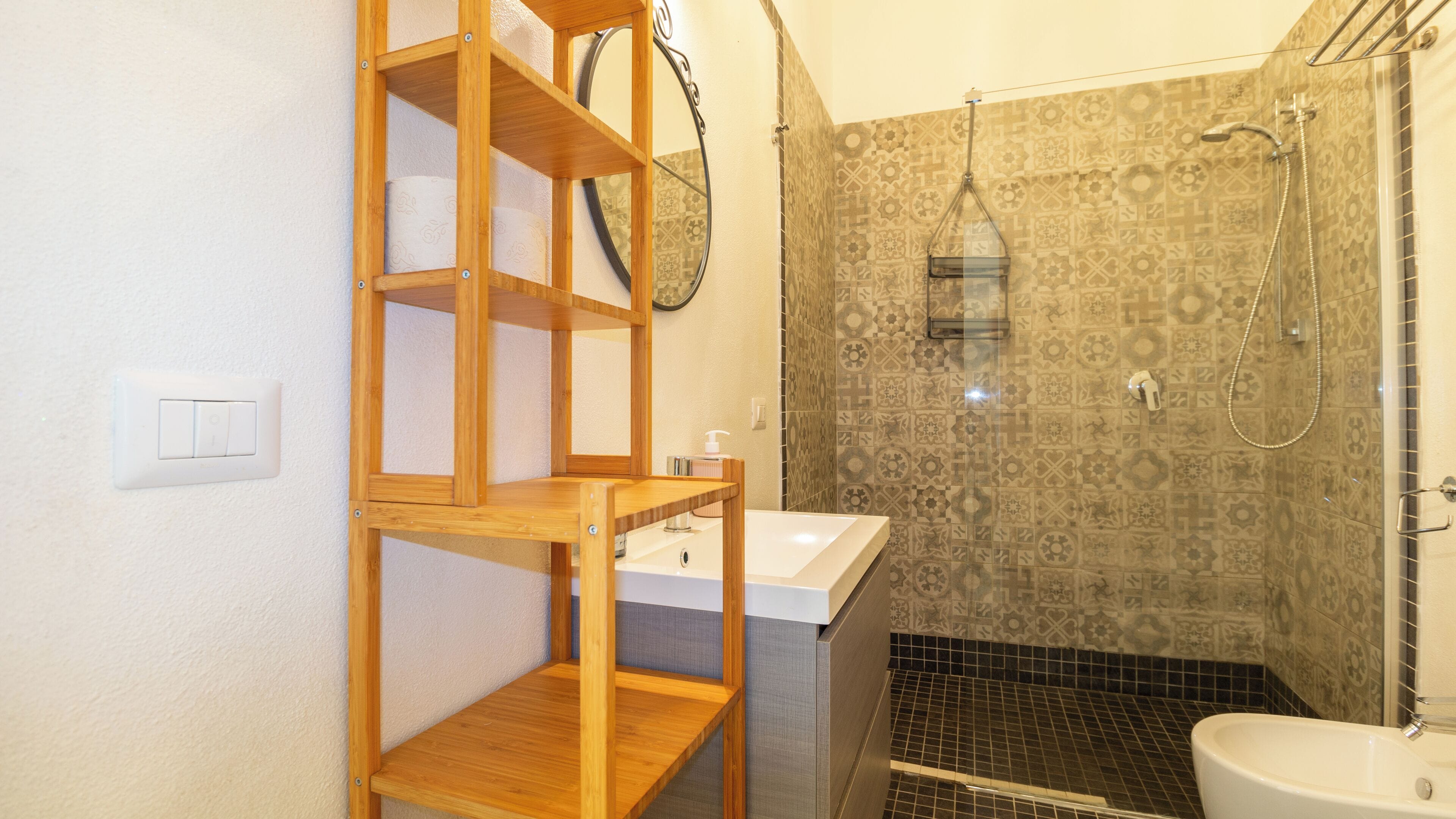 A bathroom with a wooden shelf unit, a sink, a toilet, and a shower area with patterned tiles.