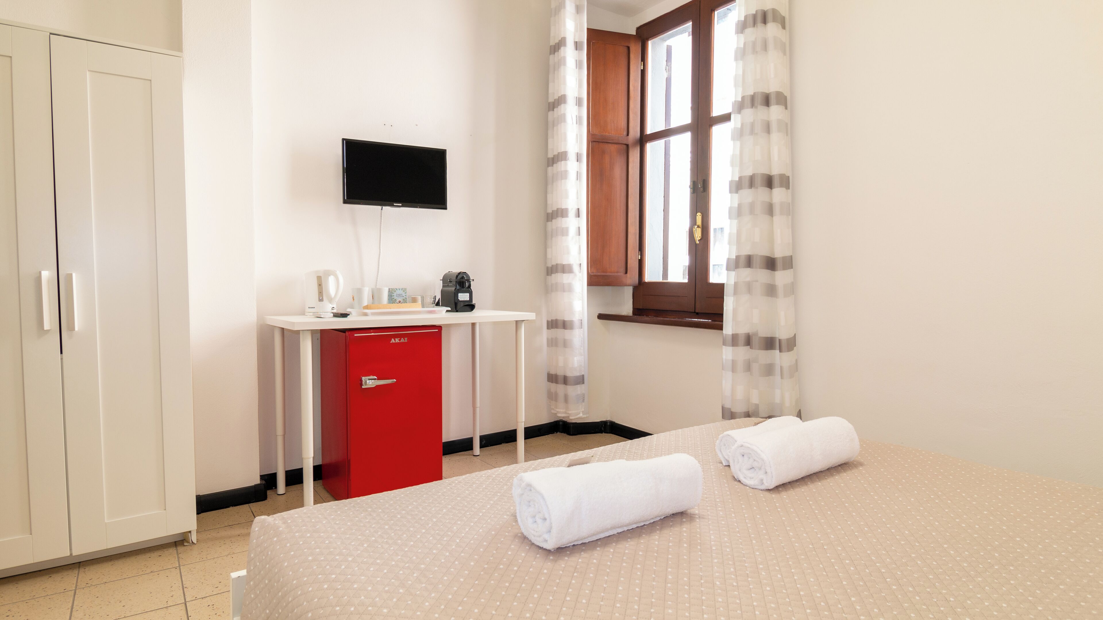 Una camera d'albergo con un letto, una scrivania, un minibar rosso, una televisione e una finestra con tende.