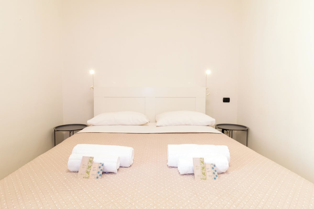 Un lit bien fait, avec du linge de lit et des serviettes blanches, deux tables de chevet et un luminaire fixé au mur.