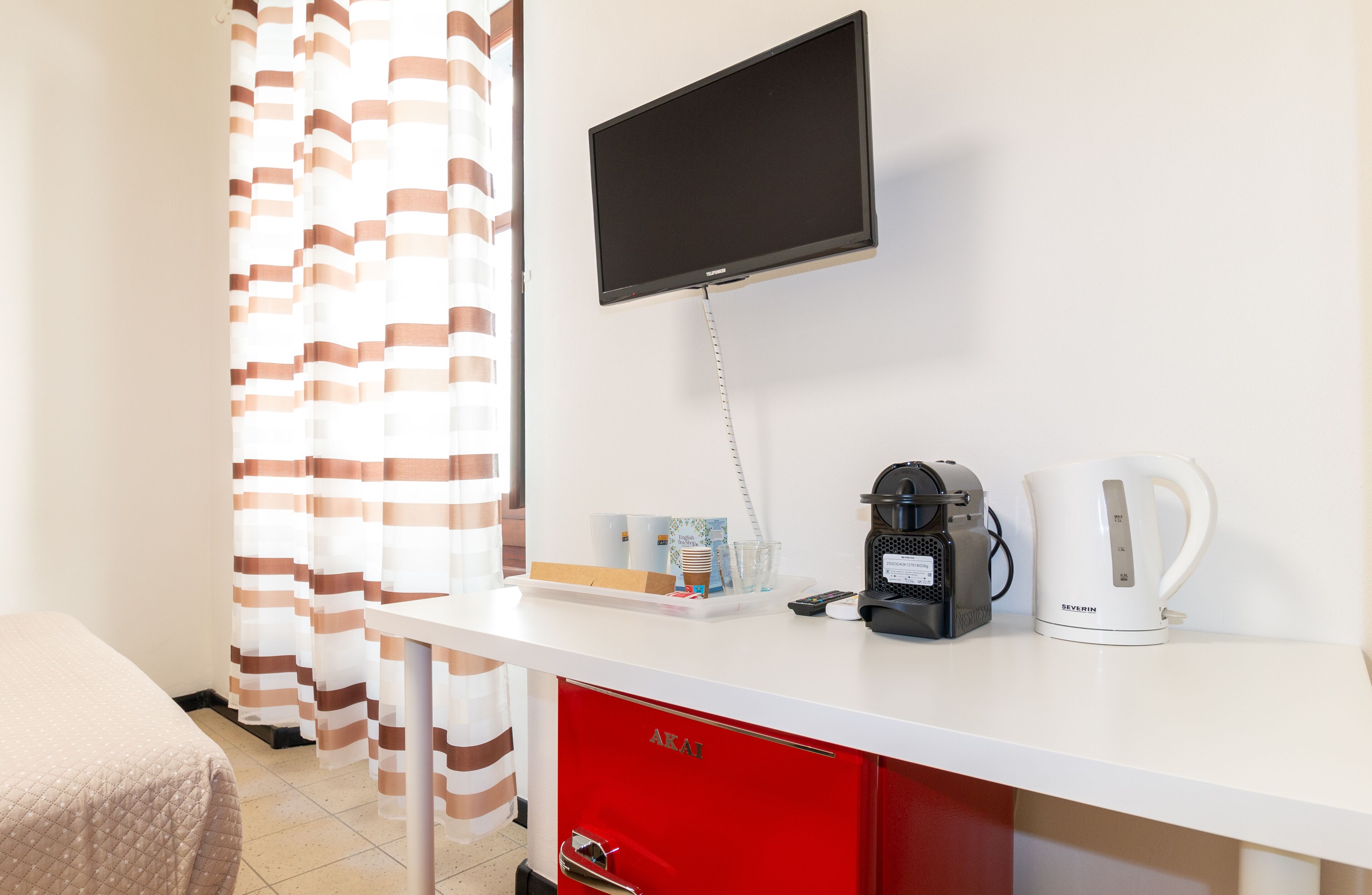 A hotel room with a white desk, a red drawer, a television mounted on the wall, a coffee maker, and a kettle.