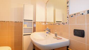A bathroom with a white sink, a mirror, and a toilet. There are shelves above the sink and a wall socket on the right.