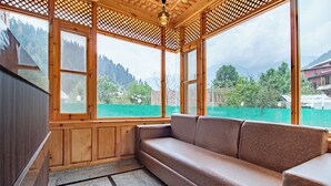 A wooden room with a cushioned bench, large windows, and a view of a green fence and mountains. - Bombay Residency (Pahalgam)