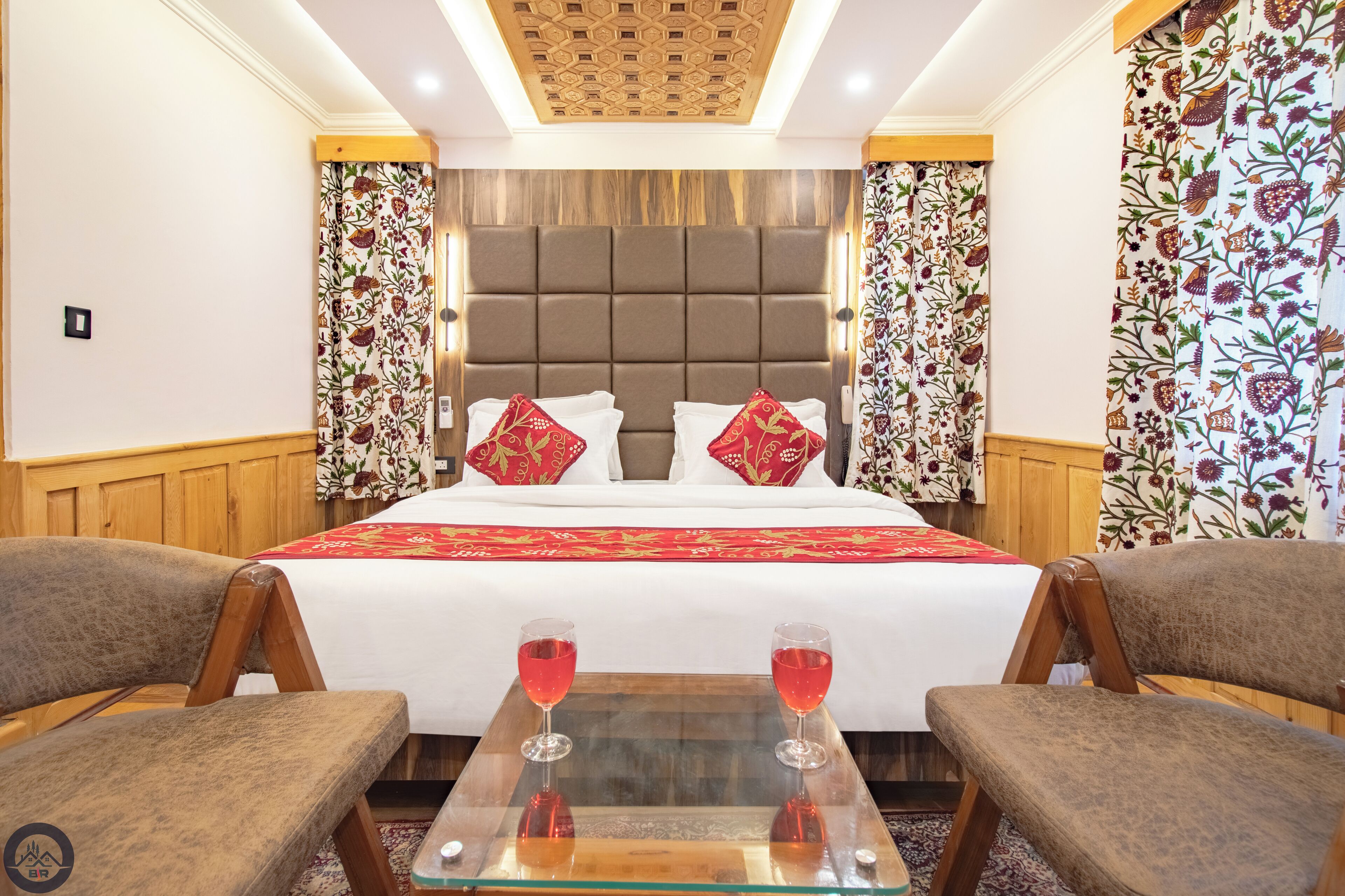 A hotel room with a large bed, two red pillows, a glass coffee table with two glasses of red drink, and wooden chairs.