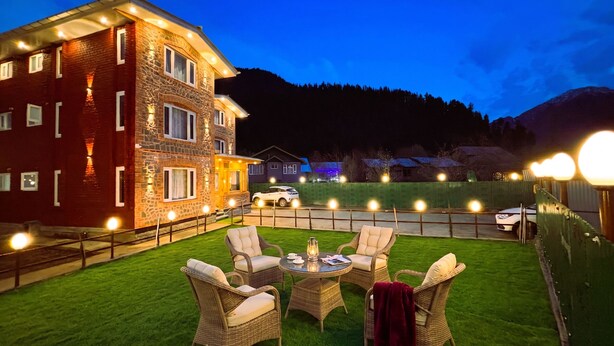 A well-lit outdoor area with a dining table and chairs, a stone building with multiple windows, and a view of a mountain in the background. - Bombay Residency (Pahalgam)
