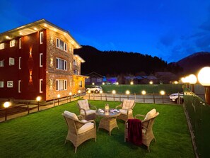 A well-lit outdoor area with a dining table and chairs, a stone building with multiple windows, and a view of a mountain in the background. - Bombay Residency (Pahalgam)