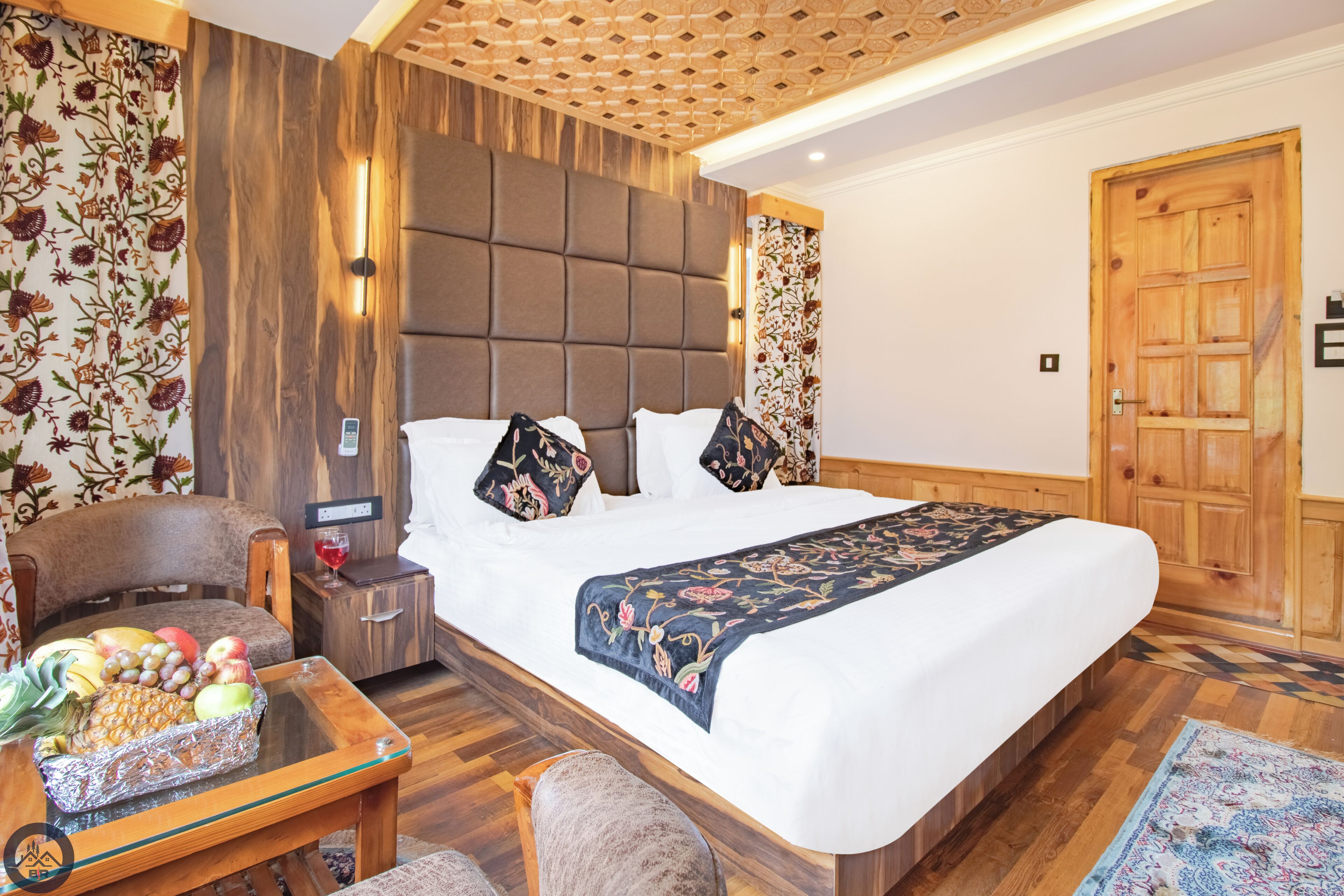 A hotel room with a large bed, a wooden headboard, a small table with fruit, a chair, and a wooden door.