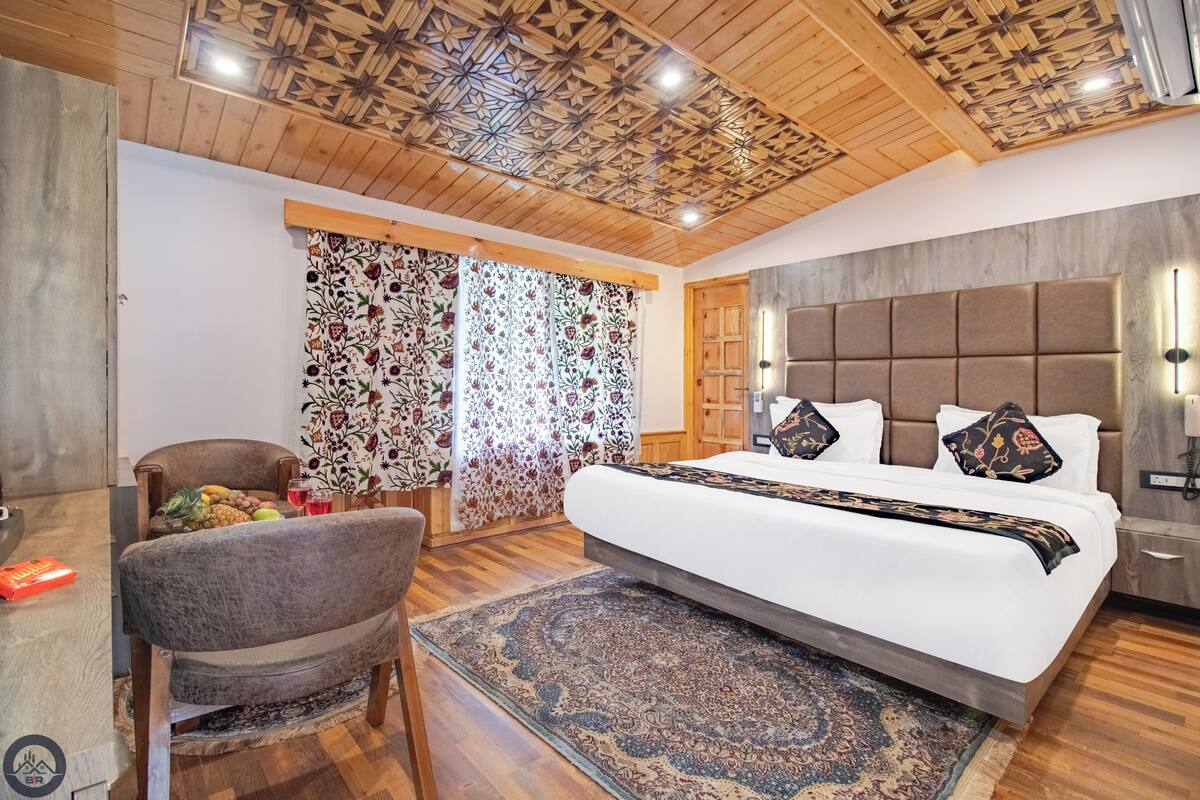 A bedroom with a large bed, a wooden ceiling, a chair, a table with fruits, and a patterned rug.