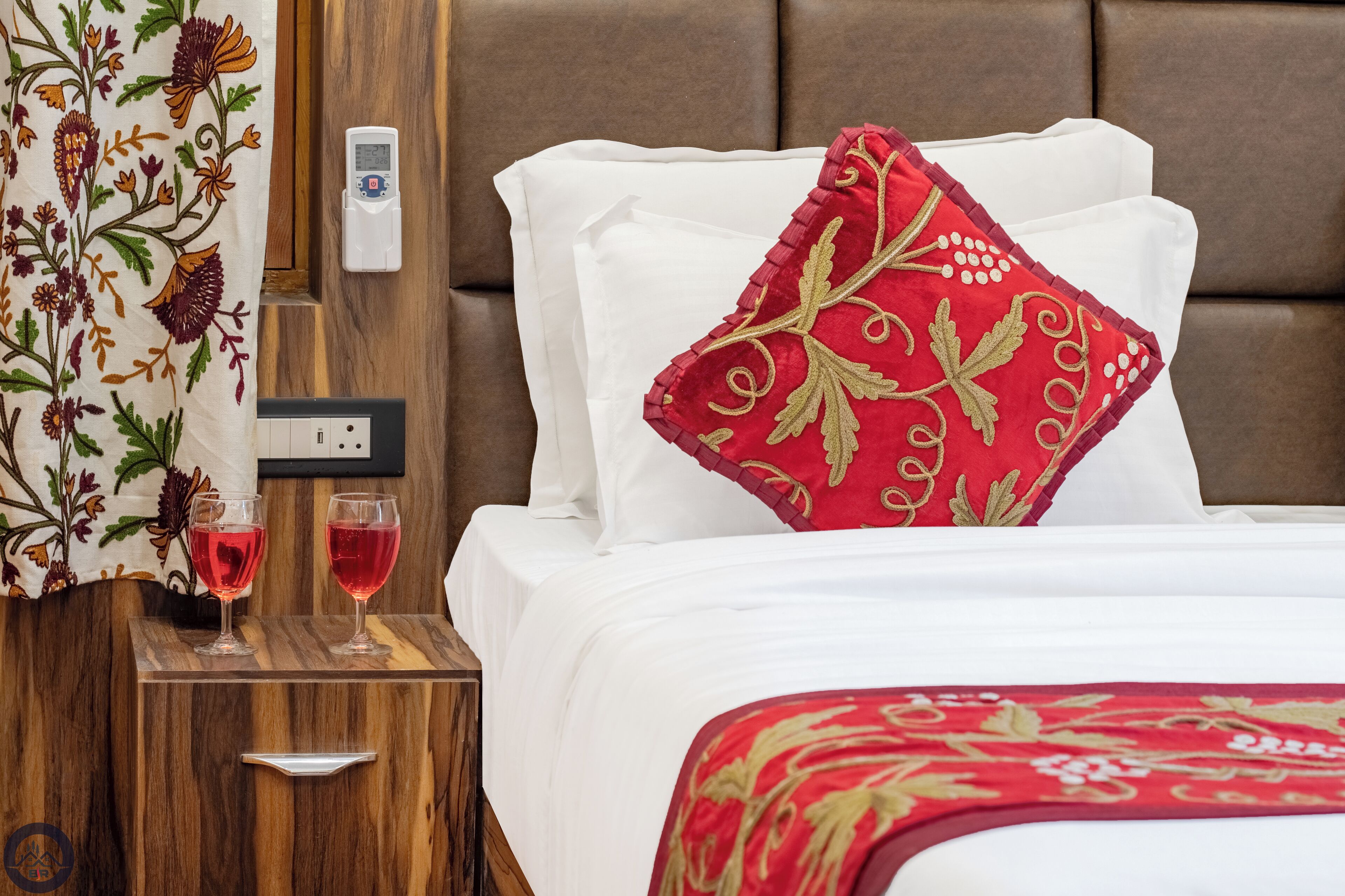 Una cama bien hecha con sábanas blancas y una almohada decorativa roja, una mesita de noche de madera con dos copas de vino tinto, y una cortina con estampado floral.