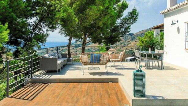 Eine Terrasse mit Gartenmöbeln, Blick auf das Meer und ein weißes Gebäude mit Schornstein.