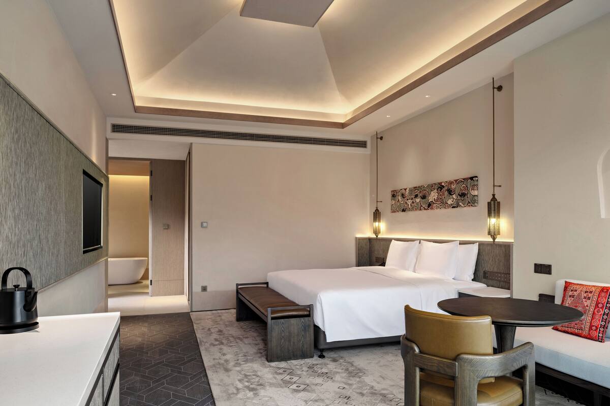 A modern hotel room with a large bed, a desk, a chair, a sofa, and a bathroom visible in the background.