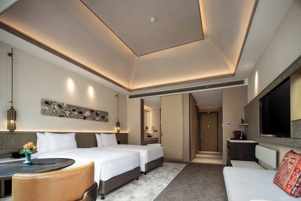 A modern hotel room with two beds, a sofa, a dining table, and a TV.