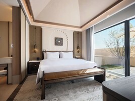 Una habitación de hotel moderna con una cama grande, mesitas de noche, un banco y un ventanal con vistas al exterior.