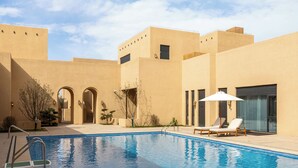A swimming pool with lounge chairs and a covered seating area, surrounded by a building with arched doorways and a flat roof. - Wanda Vista Ganman Dunhuang (Jiuquan)