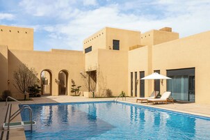 Uma piscina com espreguiçadeiras e área de estar coberta, cercada por um edifício com arcadas e telhado plano.