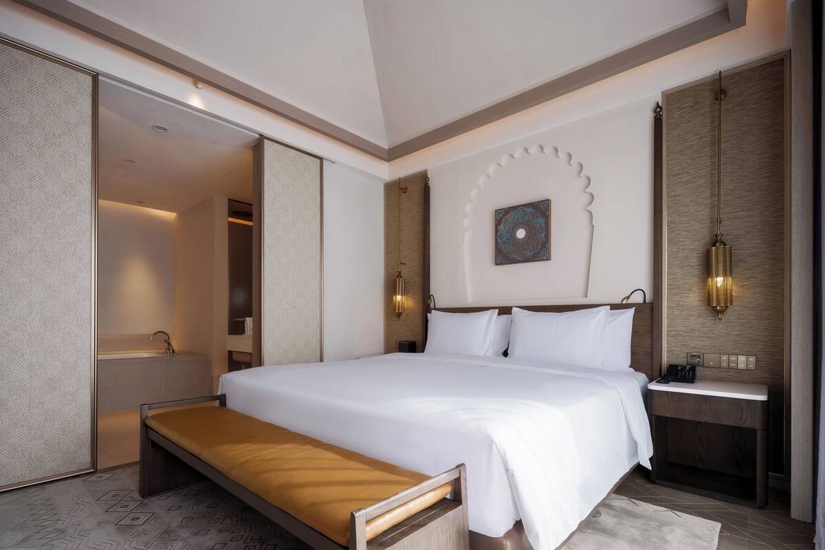 A modern hotel room with a large bed, a bedside table, a built-in bench, a bathroom with a sink, and a decorative wall piece.
