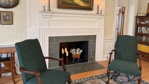 A room with a fireplace, two green upholstered chairs, a bookshelf, and a framed painting above the fireplace. - Stay 3 and play 4 free!!! (Dandridge)