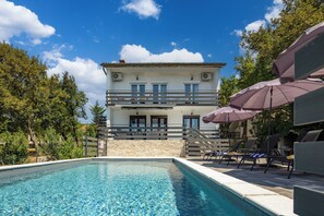 A pool area with lounge chairs and umbrellas, a stone wall, and a two-story building with balconies in the background. - Villa Natali With Pool And Sea View (Kraljevica)