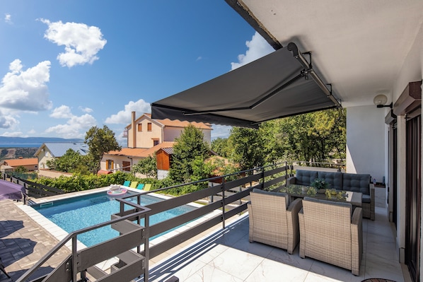A rooftop pool area with a view of houses and trees, a covered seating area, and a poolside table.