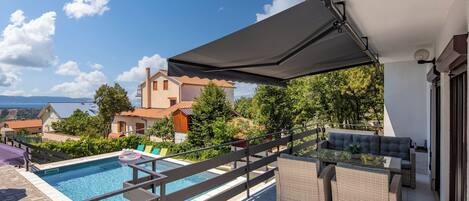 A rooftop pool area with a view of houses and trees, a covered seating area, and a poolside table.
