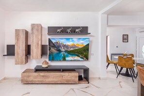 A modern living room with a large flat-screen TV mounted on the wall, wooden shelves, a wooden bench, and a dining area with a table and chairs. - Villa Natali With Pool And Sea View (Kraljevica)