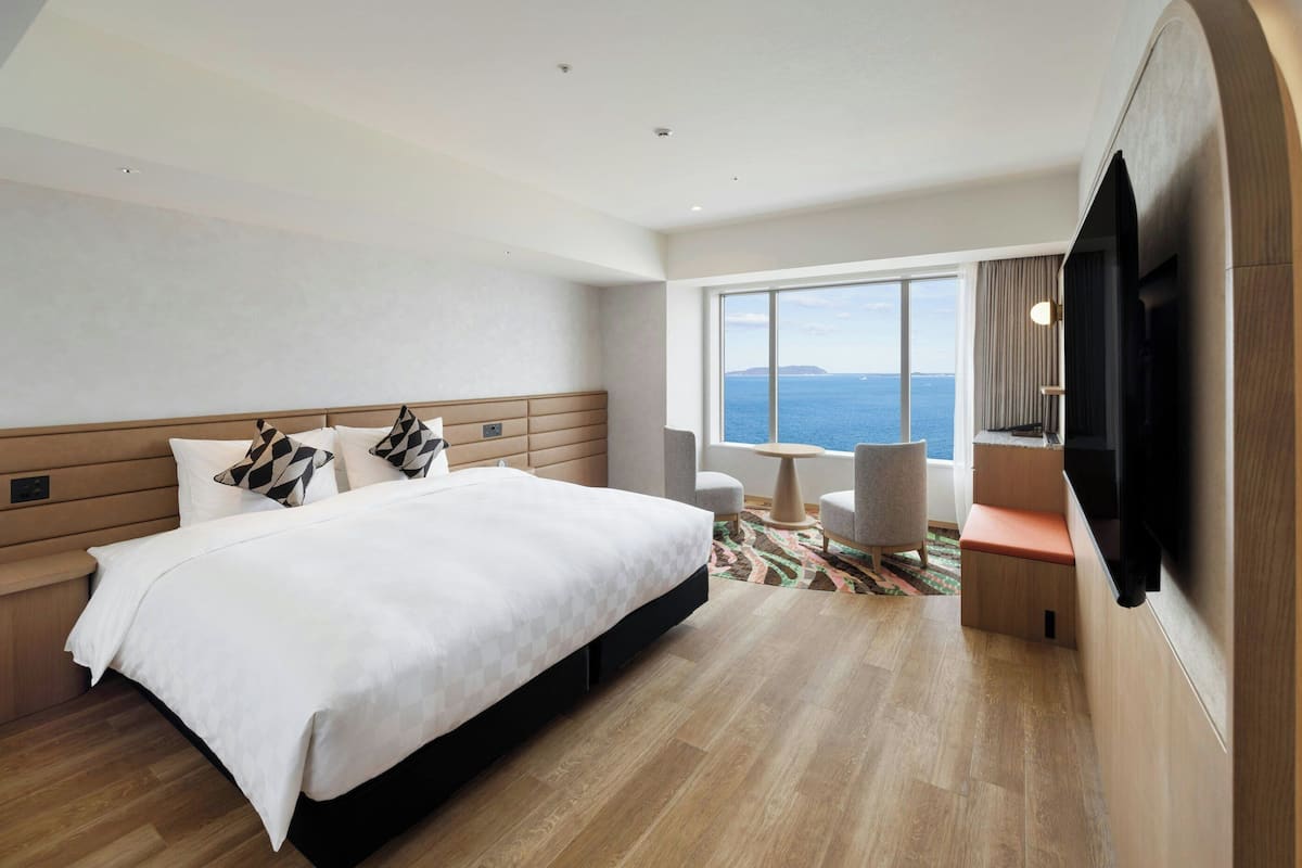 Ein modernes Hotelzimmer mit einem großen Bett, einem Fernseher, einer Sitzecke mit Meerblick und einem großen Fenster.