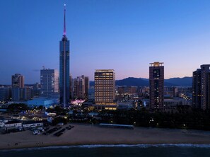 A coastal cityscape with high-rise buildings, including a prominent skyscraper with a spire, and a beach area with a parking lot and a small building.