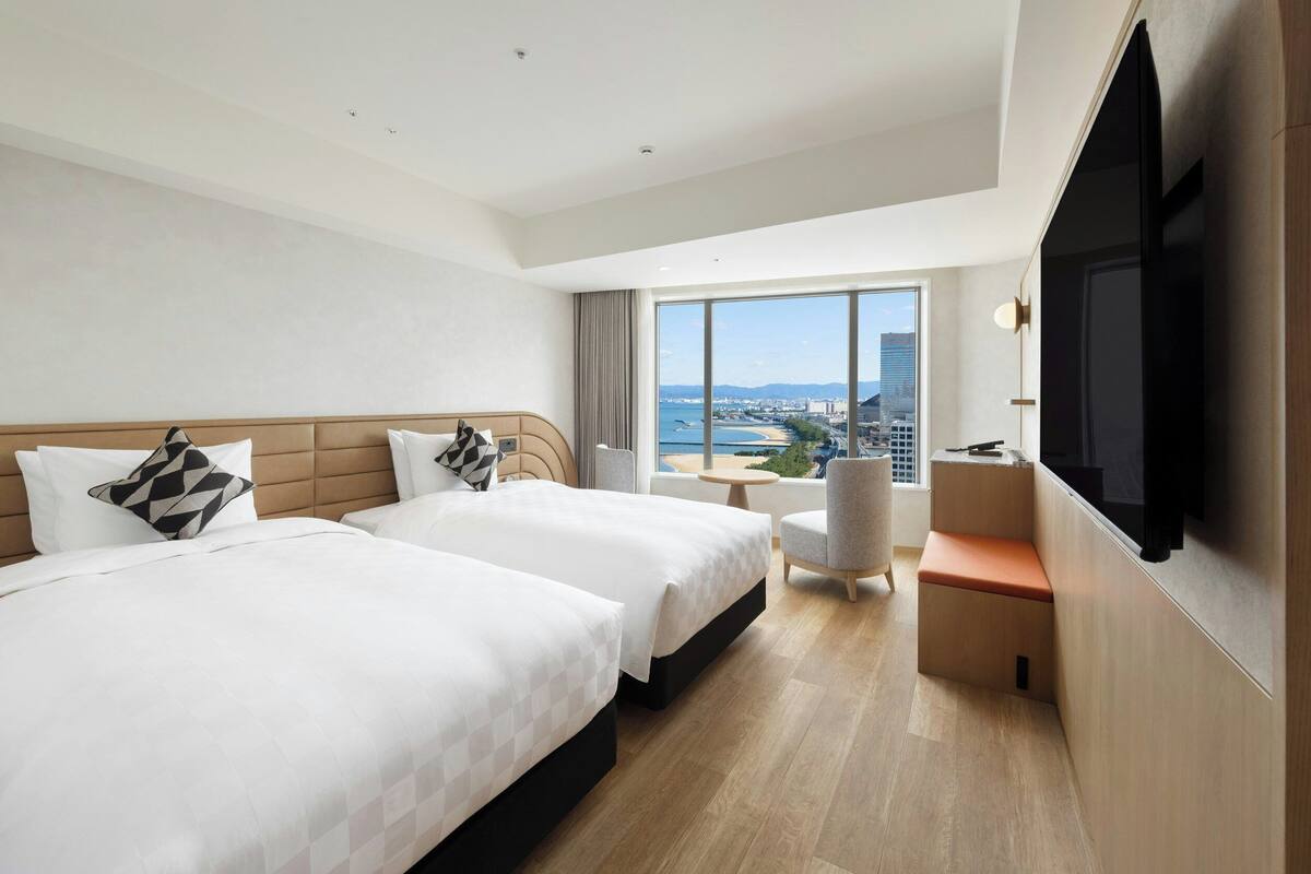Ein Hotelzimmer mit zwei Betten, einem Schreibtisch, einem Sessel, einem Fernseher und einem großen Fenster mit Blick auf die Stadt.