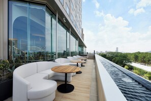 A rooftop terrace with white sofas and round wooden tables, overlooking a cityscape and greenery.