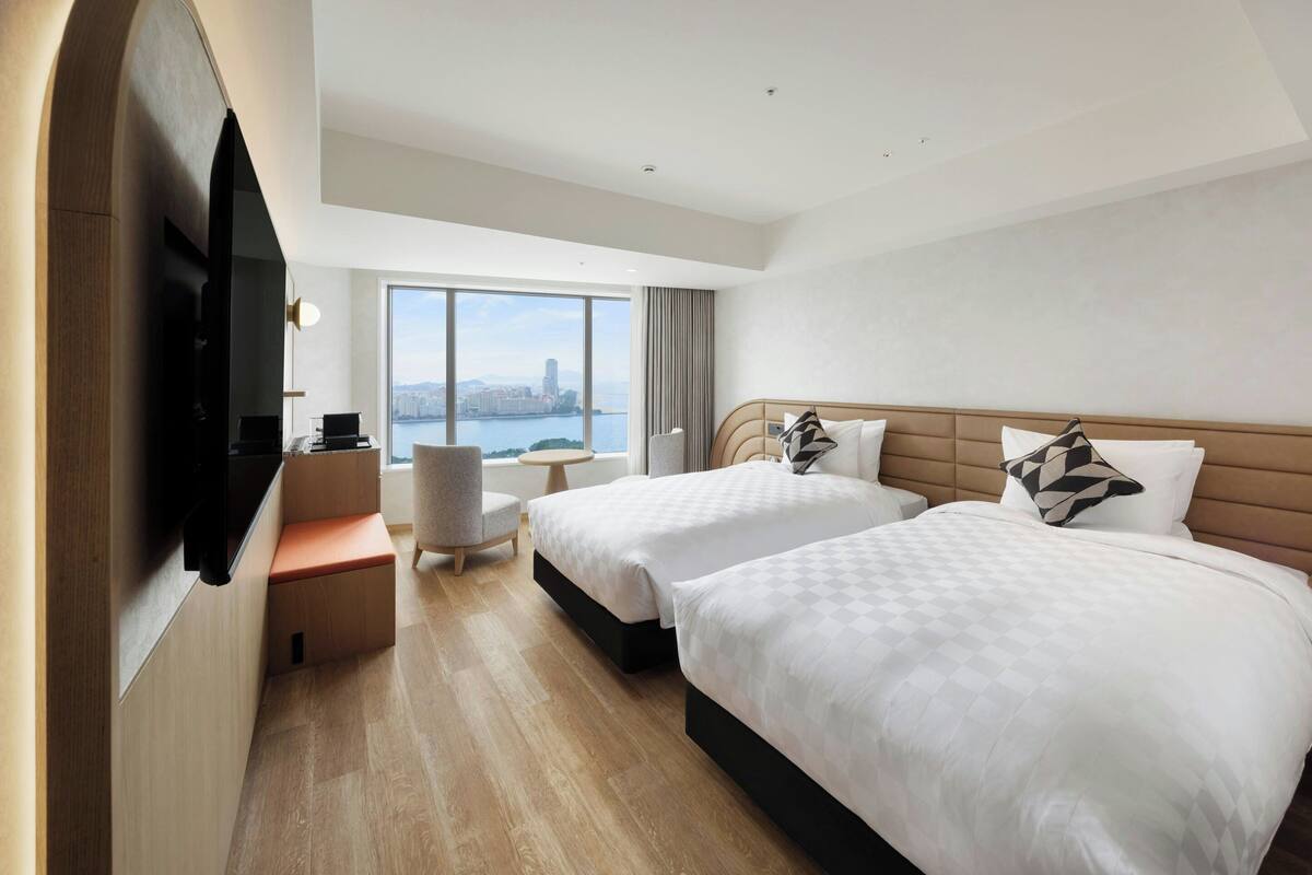 Ein Hotelzimmer mit zwei Betten, einem Fernseher, einem Schreibtisch und Blick auf eine Stadtlandschaft.