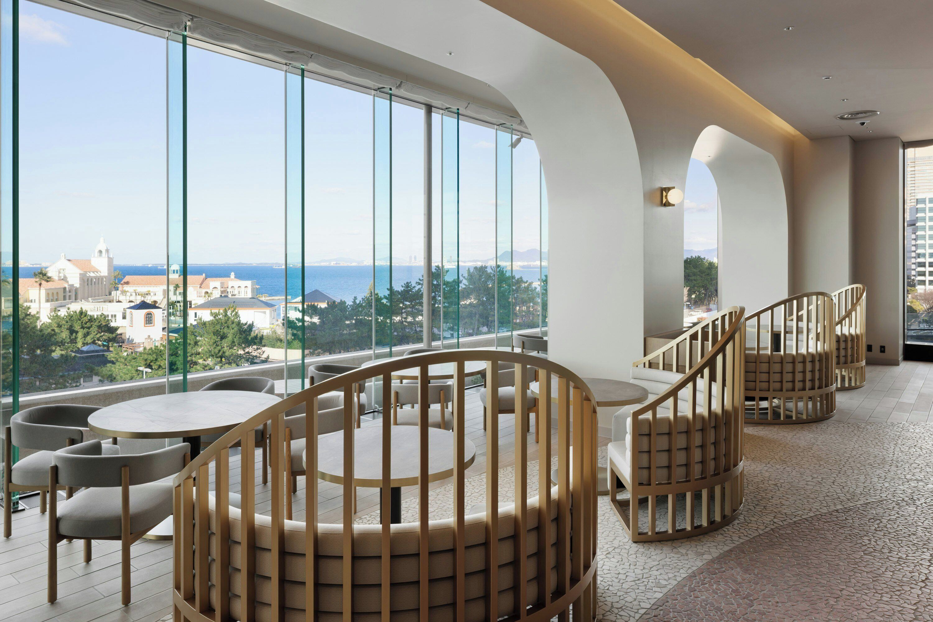 A modern interior with round tables and chairs, large windows with a view of the sea and buildings, and a curved ceiling.