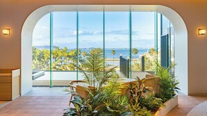 A modern interior with a large arched window, a seating area with plants, and a view of the sea. - Fukuoka Prince Hotel Momochihama (Fukuoka)