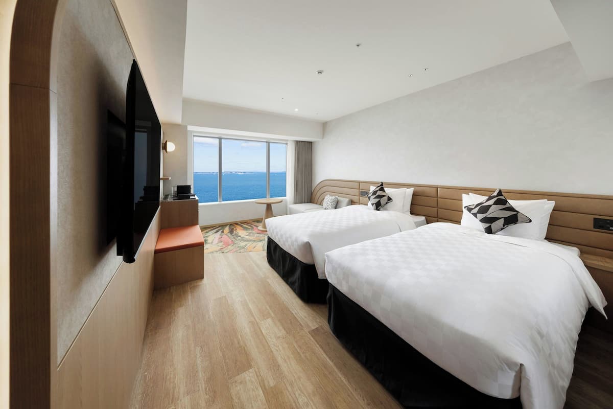 Ein Hotelzimmer mit zwei Betten, einem an der Wand montierten Fernseher und einem großen Fenster mit Blick auf das Meer.