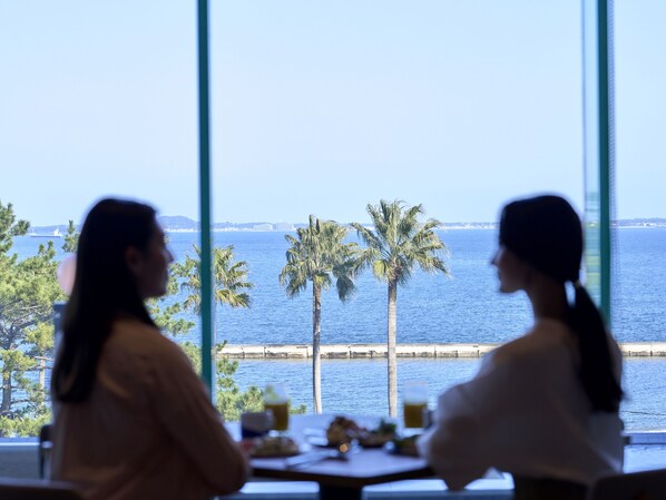 Dos personas sentadas en una mesa con vista al mar y palmeras a través de amplios ventanales.