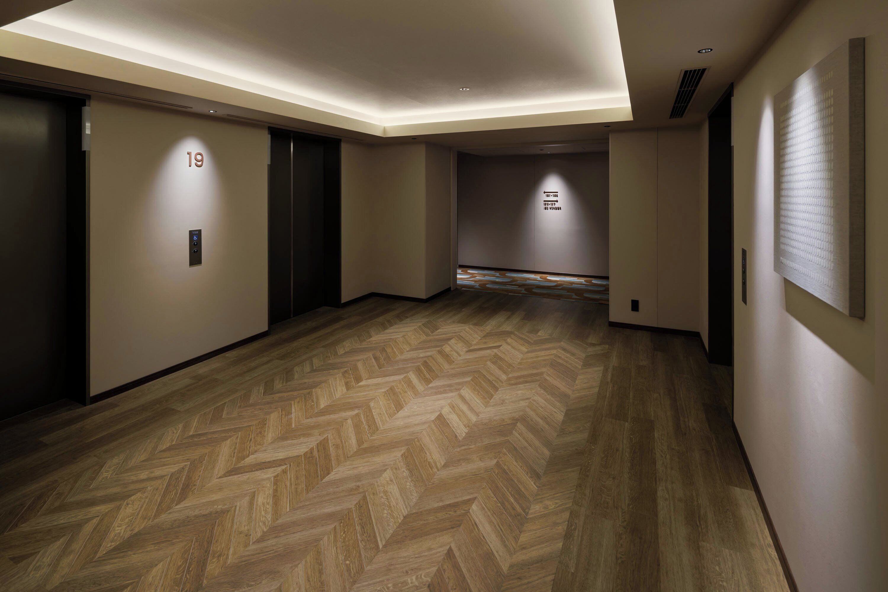 A hallway with a herringbone floor, a wall-mounted elevator control panel, and a door with the number 19.