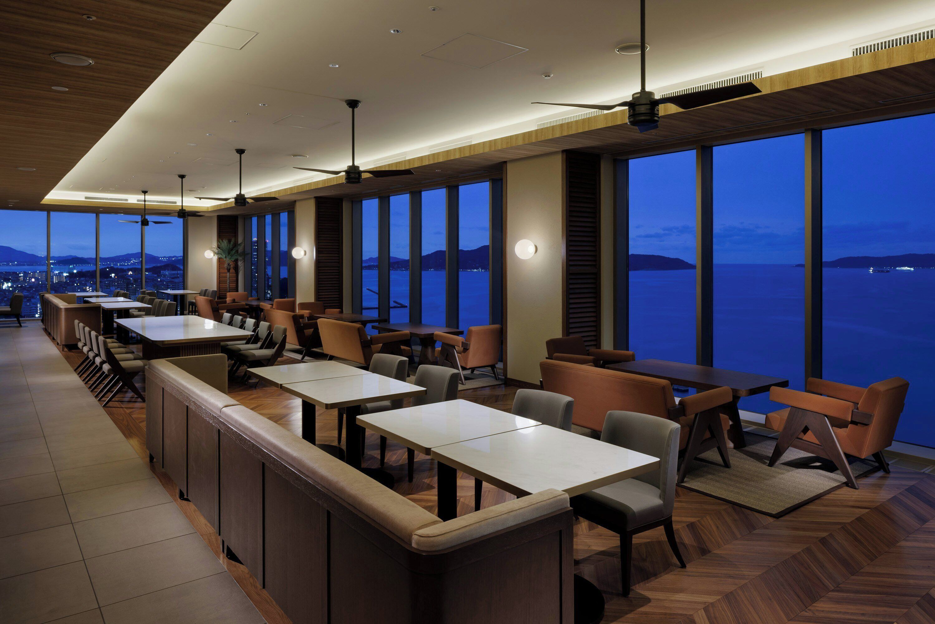 Un ristorante moderno con un lungo bancone, diversi tavoli e sedie, ampie finestre con vista sul mare e sulla città.