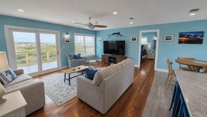 A spacious living room with a large sofa, a coffee table, and a television. There is a balcony with a view of the landscape.