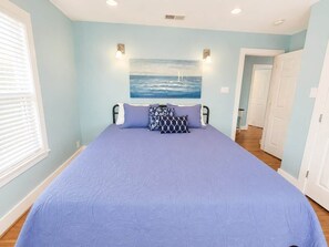 Un dormitorio con una cama grande, un cuadro de un paisaje marino y un pasillo que lleva a una puerta.