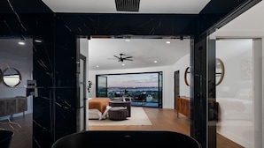 A modern bathroom with a large black bathtub, a ceiling fan, and a view of a cityscape through large windows.