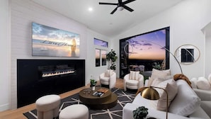 A modern living room with a fireplace, comfortable seating, and large windows offering a view of the sunset.