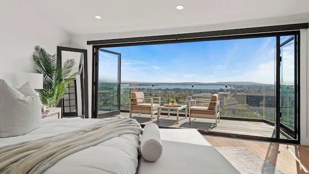 A modern bedroom with a large bed, a balcony with seating, and a city view.
