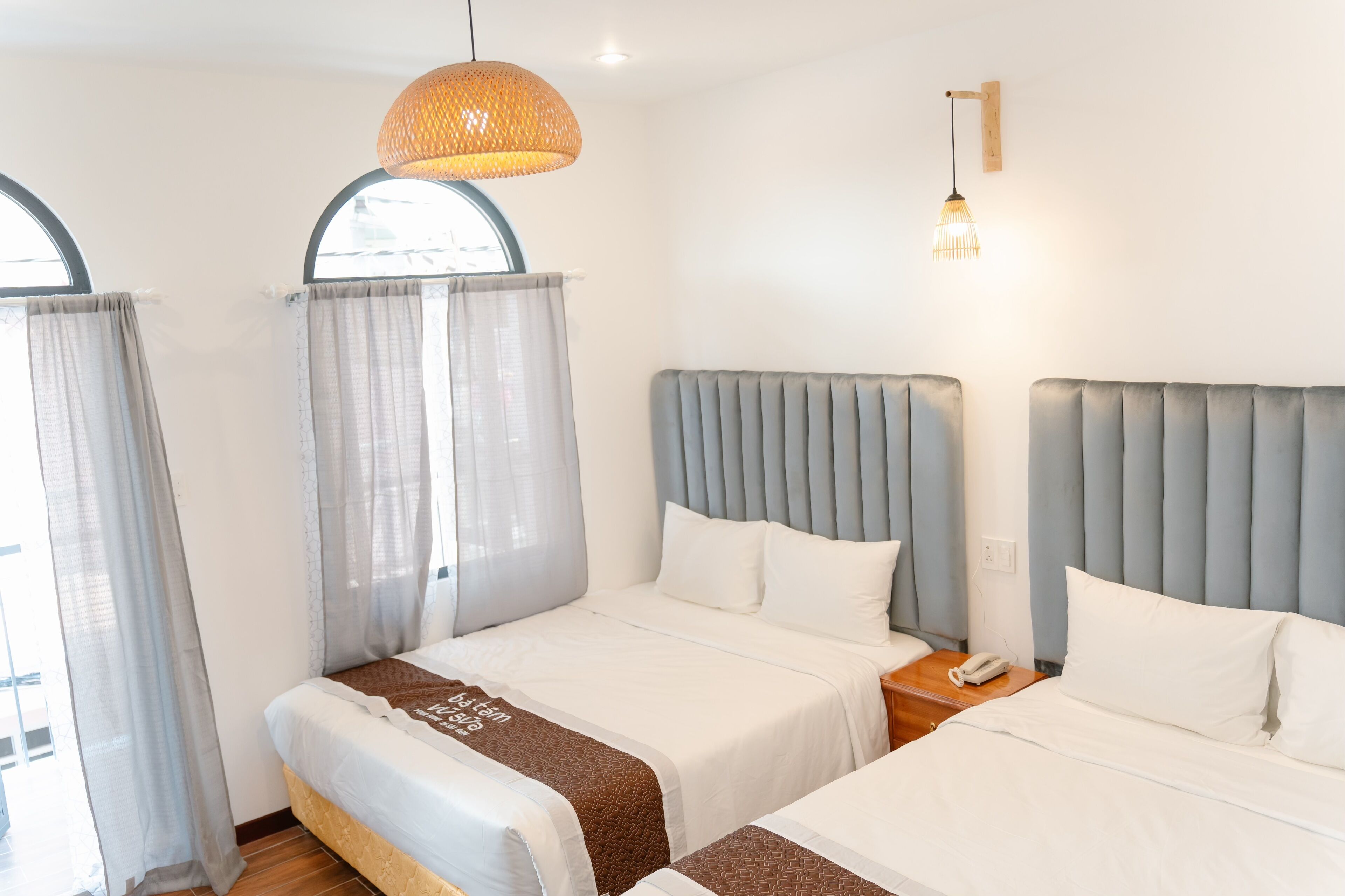 A hotel room with two beds, a large window with curtains, a hanging light fixture, and a bedside table with a phone.