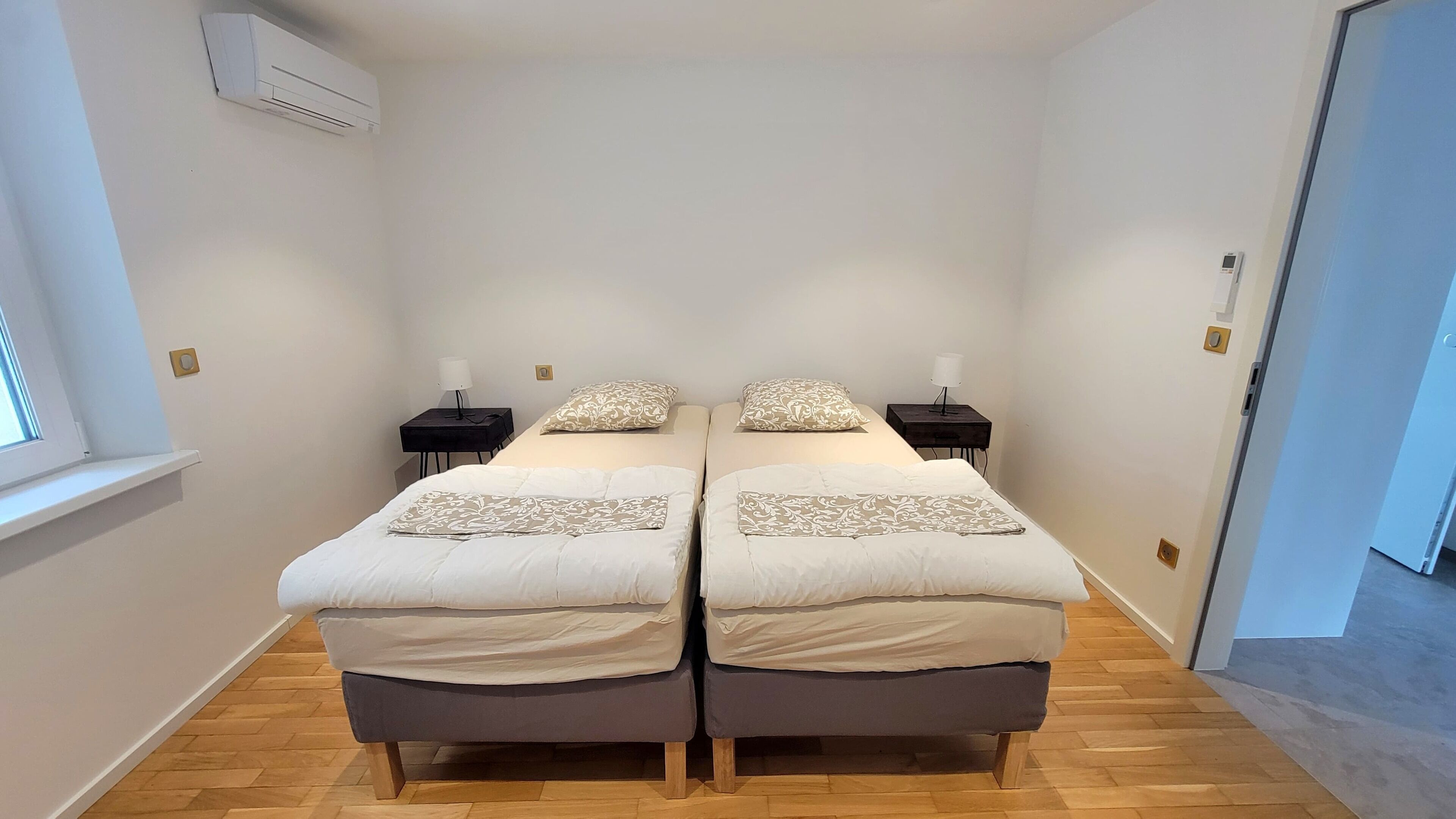A double room with two beds, each with a bedside table and lamp, a window, and an air conditioning unit on the wall.