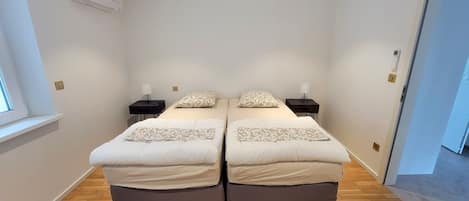 A double room with two beds, each with a bedside table and lamp, a window, and an air conditioning unit on the wall.