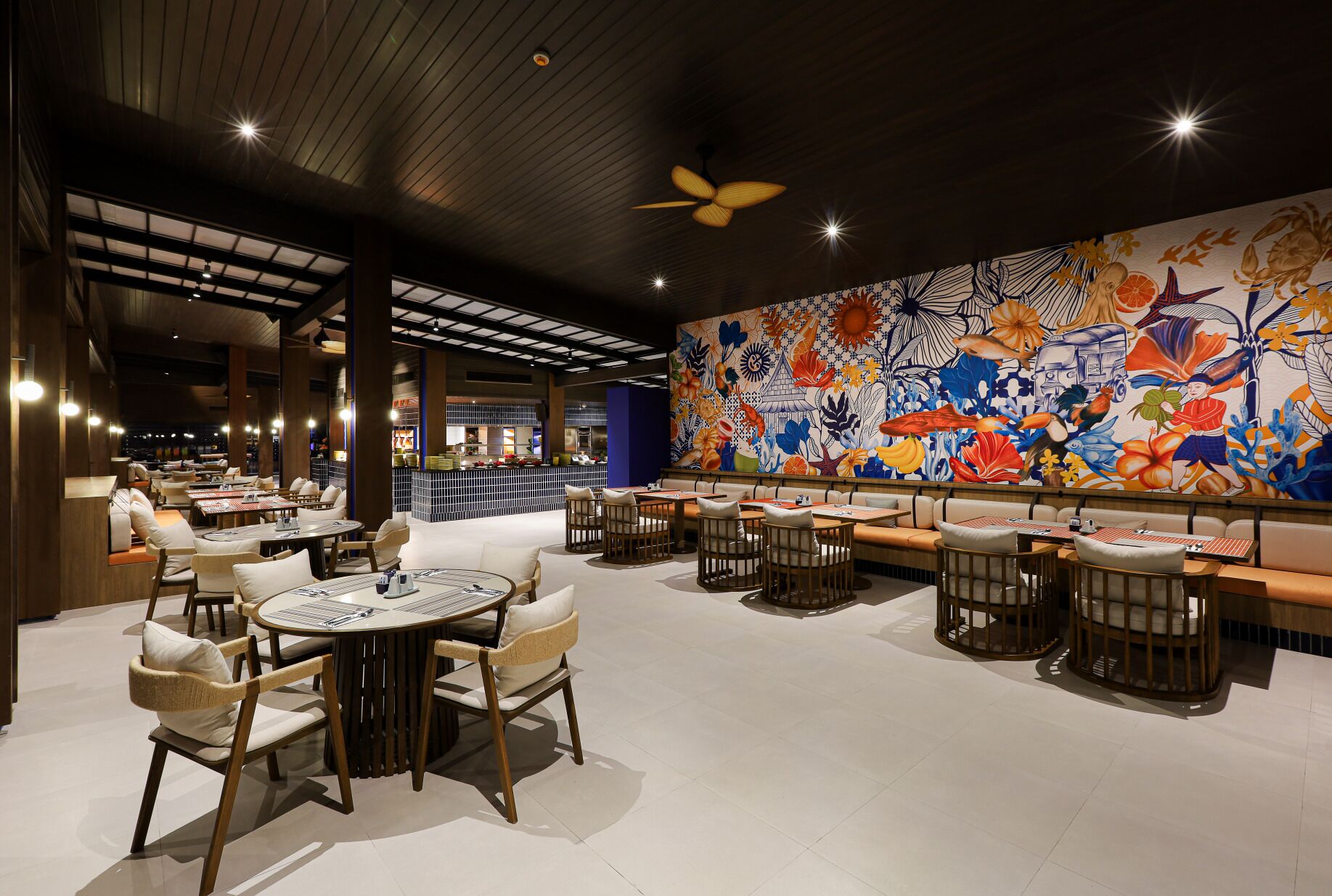 Un restaurant avec des tables rondes et des banquettes, une grande fresque murale aux motifs tropicaux et floraux, et un ventilateur de plafond.